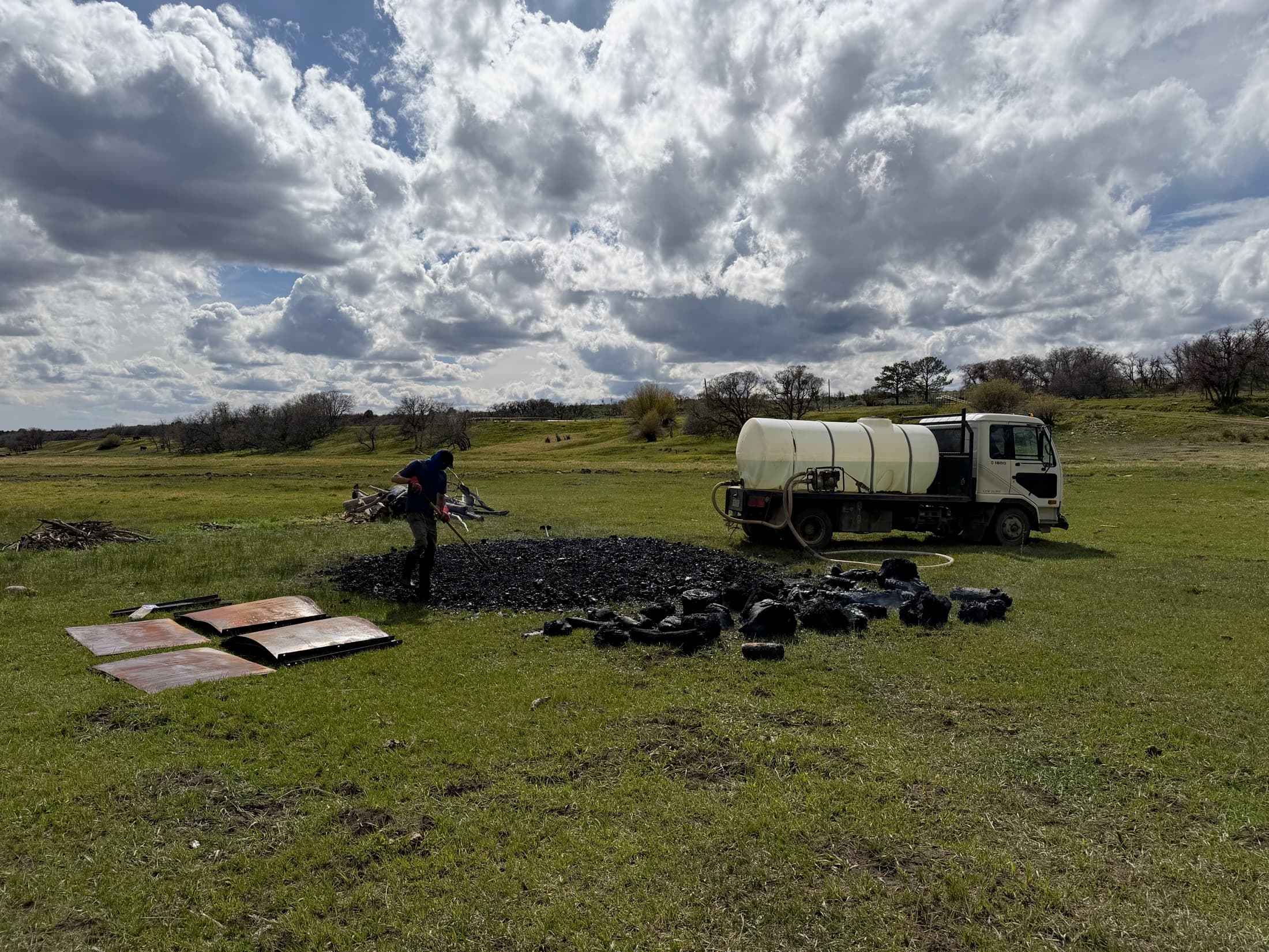 A Gardens of Fire crew member raking out a fresh biochar harvest beside the water truck under a wide Four Corners sky