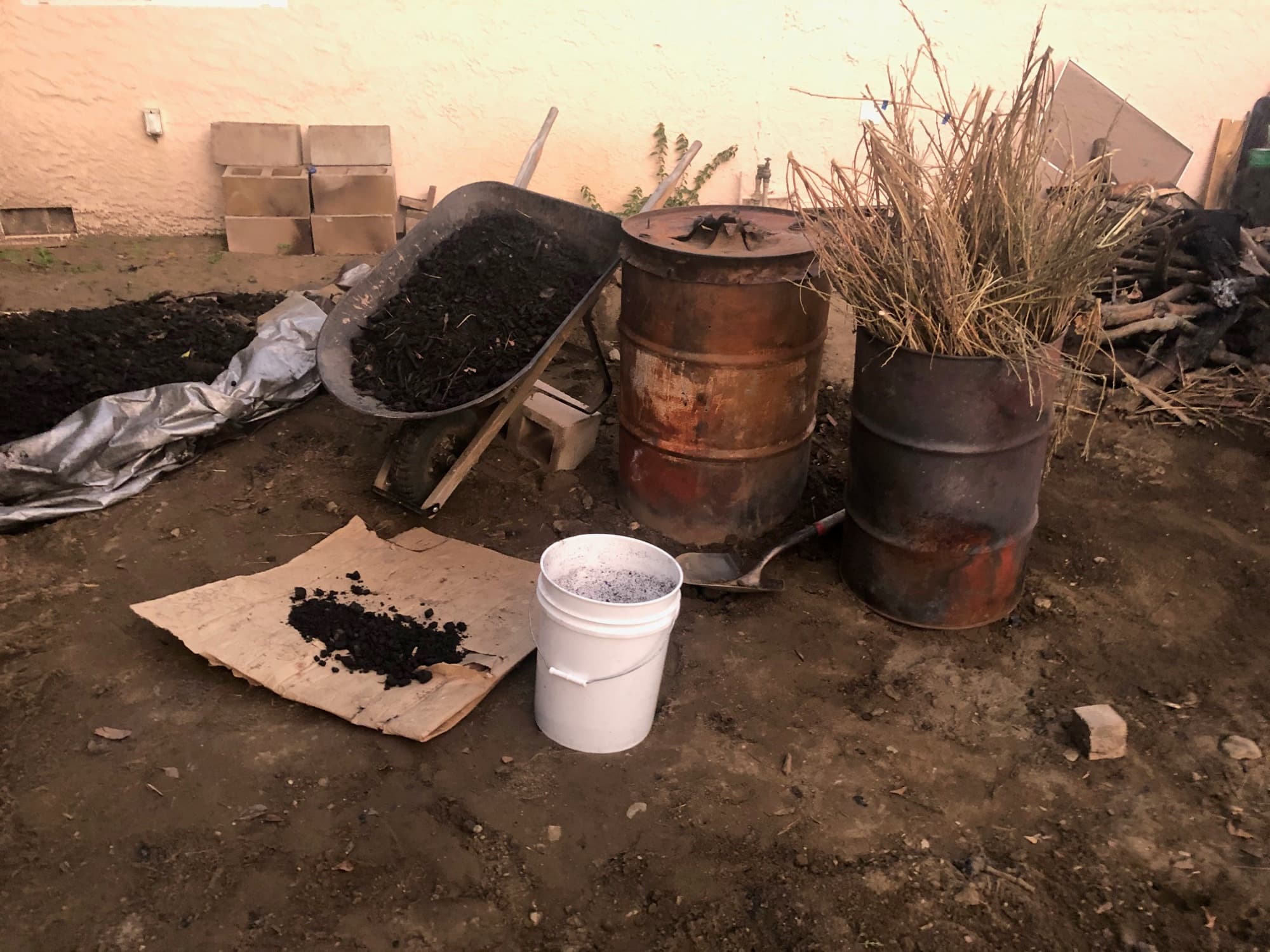 A wheelbarrow of fresh char beside two barrel kilns and a bucket of the first biochar